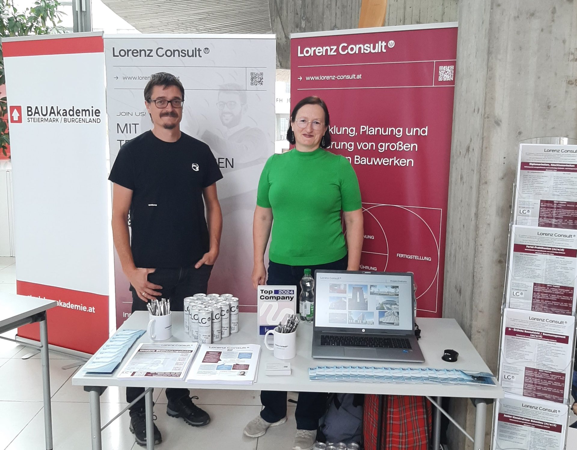 Two team members at the FH JOANNEUM info day booth, surrounded by giveaways and informational materials for visitors.